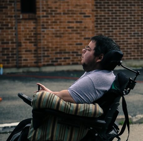 A photo of filmmaker, Dominick Evans, a wheelchair user, sitting on set. There is a brick wall behind him, since he is outdoors. He is looking towards something off-camera with an intent look. It is a view of his profile.